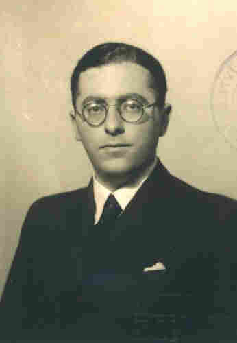 A portrait photo of a man in a suit and round-framed glasses. You can see his face and chest. He has short hair. 