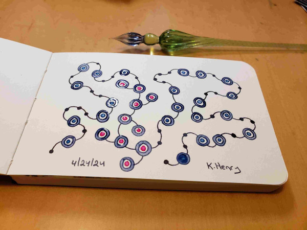 Hand drawn generative art in ink on an open page of my sketchbook. Abstract pattern looks a bit like beads on a string with knots. My glass dipping pen is next to my sketchbook.