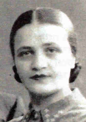 Black and white photograph of a mature woman's face. She has dark hair with a centre parting pinned back. She has painted lips. A pattern of light crosses can be seen on her collar.