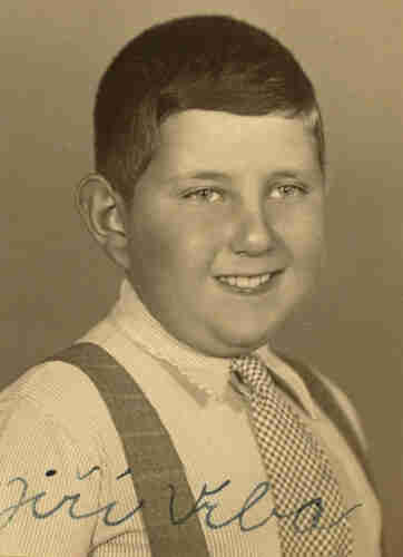 Portrait photograph of a young teenage boy. He is smiling and has his head turned slightly so that his right ear is visible. He has short dark hair. He is wearing a fine checked tie, a very thin striped shirt and darker braces.