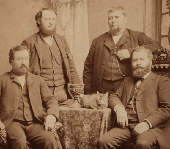 Sepia toned black and white photo of four stout gentlemen posing for a group portrait, two are seated in the front, two standing in the back, with a rug-covered small table between them all. On the table is an ornate chalice and a shorthaired tabby cat.