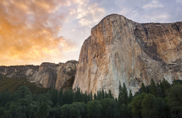 The stunning El Capitan granite monolith at Yosemite National Park, California, USA. Photo taken at sunset from the valley floor, looking upward at the sheer rock face against a colorful partly cloudy sky.