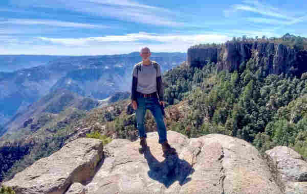 Me, looking toward the camera, standing near the edge of a precipice facing out over Mexico's Copper Canyon. Behind me is an expansive vista of a spectacular broad and deep canyon, some trees, and a blue sky with a few clouds.