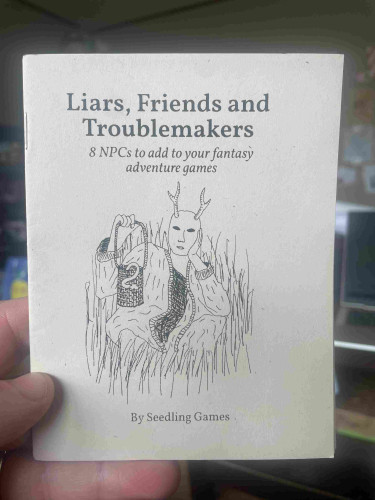 A hand holding a book titled "Liars, Friends and Troublemakers - 8 NPCs to add to your fantasy adventure games" by Seedling Games, featuring a sketch of an antlered fantasy character reclining in the grass on the cover.