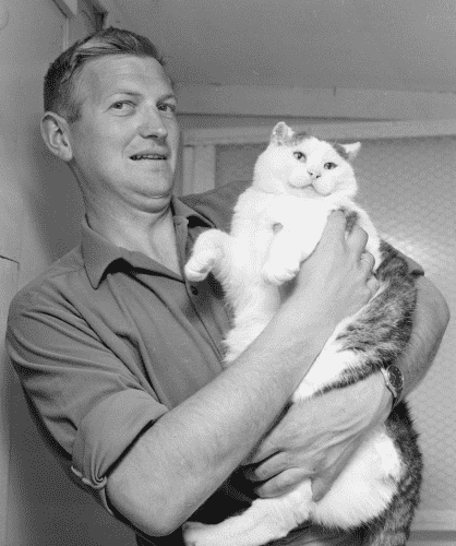 Black and white photo of a slim white man in button up shirt, showing off a chubby shorthaired white and tabby cat that he holds in his arms. The cat has tiny ears that looks like they might've been operated on at some point to partially remove the ear tissue due to frostbite or cancer, but the cat seems perfectly healthy and it has a very cute effect because it exaggerates the proportions of little round head and big chonky body. The cat also bears an adorable smug smile.
