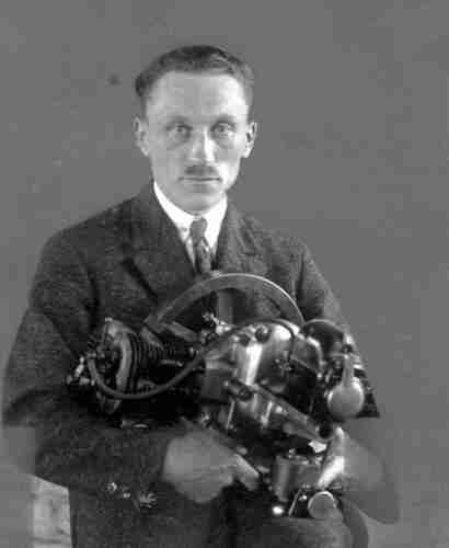 A man in a suit is holding a large mechanical structure - an engine or part of an engine. He has short hair and a moustache. 