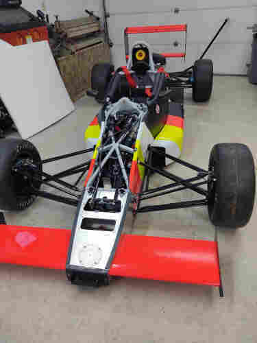 A formula racing car, in process of being rebuilt. It has pretty much brand new everything, and is almost complete. The front upper body panel is off, showing suspension and wiring that were being worked on at the time. The front fairing is bright red, and the body is striped red, yellow, white, yellow,red,and black, in order. Brand new Hoosier slicks on all four corners. Yee Haw.