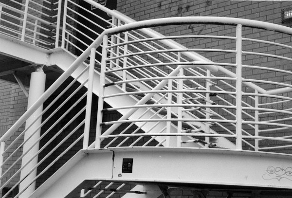 Black and white photo showing a white-painted metal fire escape as it zig-zags on the side of a building. Lots of white bars criss-crossing.