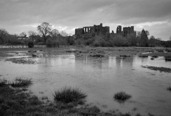Ruined castle against the horizon, reflected in the water, but with less detail as the water is not still (I think it was raining slightly). Grass at the bottom left with some bunches of reeds, a small spit of grass from mid right, otherwise most of the bottom half is water. Black and white photo.