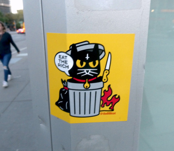 A small, angry looking but very cute black cat wearing an upside down cross on their forehead and a red collar with a bell. Kitty is holding a knife and saying, "Eat the rich" in a cartoon word bubble from within a steel trash can and wearing the lid from the can as a helmet. There's a bag of garbage behind the can, which the cat probably removed so they could use the can. There's also a small fire on the ground along the side of the trash can. 