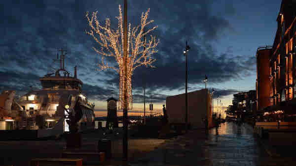 A photo of Aker Brygge at twilight. There is a ferry moored at the left of the photo, a lighted decoration of a leafless tree attached to a light post in the center of the photo, and a stone walkway leading into the distance on the right side of the photo. The sky has dark clouds in it.