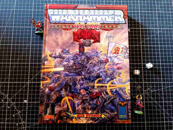 Photograph of the facsimile reprint of Warhammer 40.000 Rogue Trader with a kick ass cover with space marines shooting left and right. Next to the thick book there are two Chaos Space Marines miniatures painted by my 25 years ago and a pair of d6 dices.