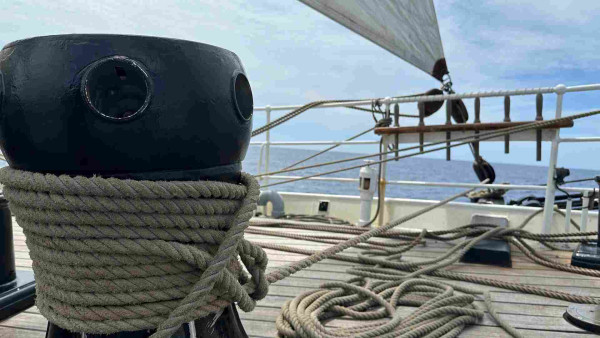 Capstan with a tugboat hitch in the foreground and the corner of a sail with two blocks almost on the deck in the background. 