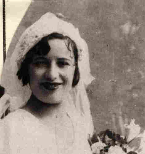A portrait photo of a young smiling woman in a wedding dress. She has semi-long dark hair. She is holding some flowers in front of her.