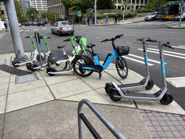 A sidewalk corner is completely filled and blocked by a collection of 5 electric scooters and 2 bikes, Lime and Bird brands. 