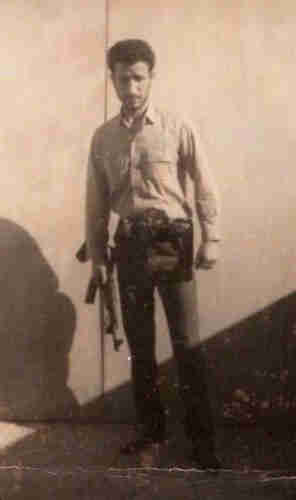 Sepia-toned photo of young man with dark hair and closely trimmed beard, wearing white long-sleeved shirt and dark pants, carrying machine gun in right hand. He is looking directly at the photographer. 