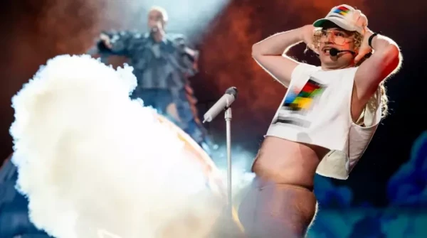 A blonde man with a curly mullet and moustache wearing a white blurred Windows 95 hat and sleeveless belly top. He leans back with his hands behind his head thrusting his crotch into the audience. A cloud of artificial smoke covers the skin coloured thong that he is wearing. An ornately denim-covered singer is in the background.  