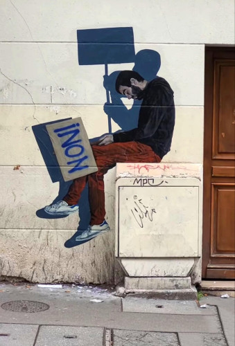 Streetartwall. The life-size, self-painted picture of a sleeping man has been stuck to a yellow house wall. The dark-haired man with a full beard sits huddled on a (real) distribution box and sleeps. He is wearing red trousers, a black sweater and sneakers. He is holding a sign with the French word "non" (English: no) written on it. Behind him is his shadow, who is not sleeping but holding up the protest sign.
Info: Notes by the artist Levalet: "In these troubled times, we can remind ourselves that each of us has the power to say no, to resist, even when the worst seems certain. Let's wake up!"