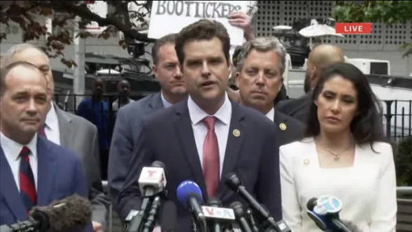 Matt Gaetz + entourage, at Trump hush money trial, in front of press microphones, with a protester holding a sign behind them that reads,

"BOOTLICKERS"