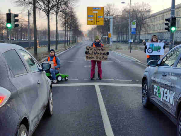 3 Menschen der letzten Generation blockierten in Leipzig eine Straße, die eine Person sitzt auf einem Kinder Spielzeug Trecker, die Person in der Mitte hält ein Schild auf dem steht, wir dürfen das, wir haben einen Traktor, die dritte Person hält ein Schild mit aufgedrucktem Trecker hoch