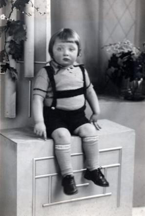 Vintage black and white photograph of a young boy seated on a small stool, wearing a striped shirt, shorts with suspenders, and knee socks. Background includes a simple curtain and potted plants.