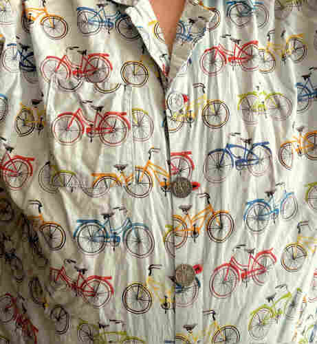 Wrinkled cotton print of classic upright bikes with chainguards, fenders, and racks. They're red, green, blue, and yellow on a light blue background. The buttons are large and metal with an intricate bas-relief of a 19th century cyclist posed next to a bike.