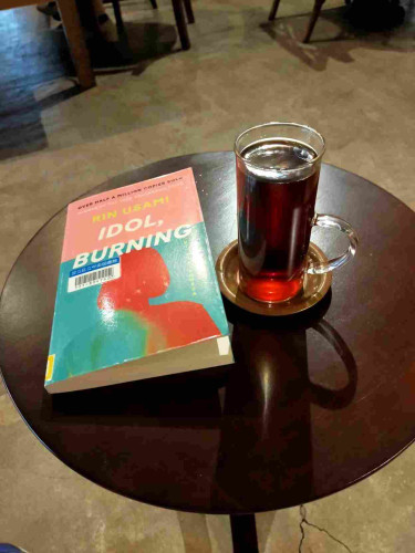The paperback library book is pink on top, turquoise on bottom with red outline of a bobbed women. To the left is a ling cylindrical glass with handle in the lower part full of black coffee on a brass coaster. All are on a round dark brown wooden table above a cement floor in a cafe