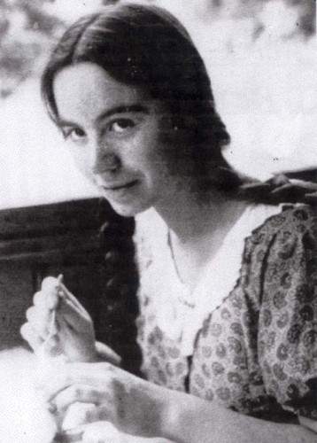 Historical photograph of a woman smiling subtly at the camera while sitting outdoors on a bench, holding an object in her hands.