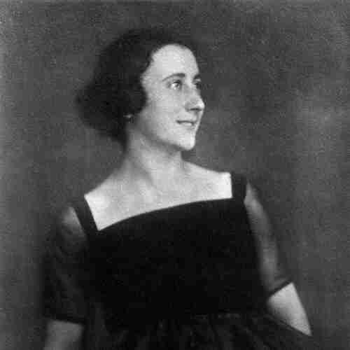 Photo of a young woman. She is wearing a strapless dress with see-through short sleeves. Her hair is dark and pinned back. Her head turned to the right side of the frame. She is smiling slightly.