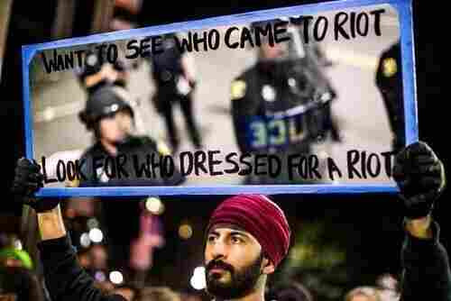 A man holds up a mirror,  reflecting police in riot gear.
Want to see who came to riot?
Look for who dressed for a riot. 