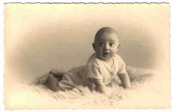 Photo in sepia. A little boy lying on his stomach on a fur blanket. He has almost no hair. His sleeves are short. He is supporting himself on his arms and looking slightly to the right of the frame. 