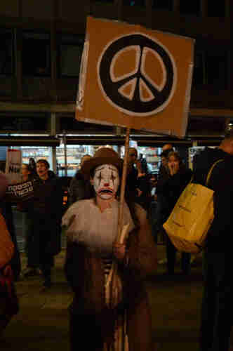A person with a painted mask on their face holding a sign with the symbol of peace on a stick.