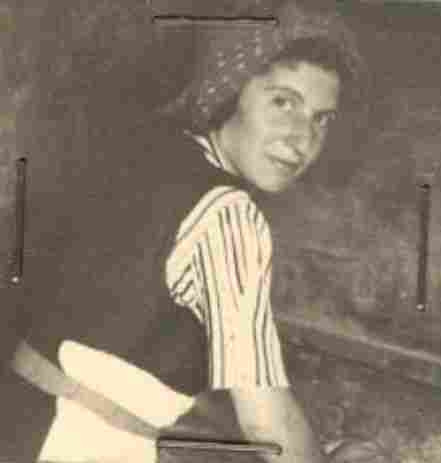A photo of a woman. She is holding her hands on some surface that is not visible. She has her hair covered with some scarf in dots. She also is wearing a vest, a short sleeve shirt in strips, and an apron. She is turning her her to the back to look at the camera.