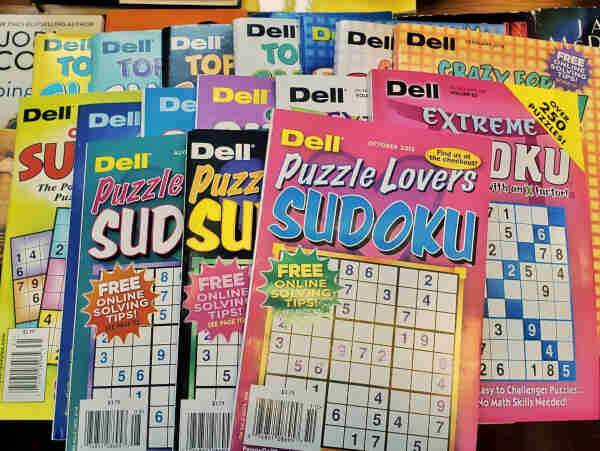 A colorful photo of 16 different Sudoku puzzle magazines from Dell publishing, fanned out on a table, such as: Crazy for Sudoku, Extreme Sudoku, Puzzle Lovers Sudoku, and more.