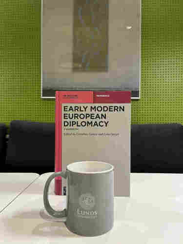 The book "Early Modern Diplomacy. A Handbook" standing on a table. In the front a grey mugg with the logo of Lund university. In the background a poster in a frame.