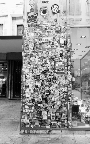 My photo with a black and white filter showing a pillar that's part of a shop front that is covered in so many different stickers that ut had created a layered effect 