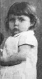 A baby girl in a short-sleeve dress. She has hair reaching her neck. She looks a bit scared and looks into the camera.