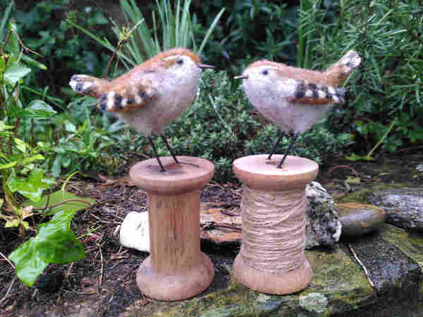 Two needlefelted wrens, life size, stood on wool cotton reels. Photo is taken outside so plants (mostly ivy) are visible behind them.