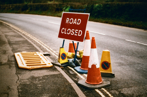 A damp road curves from bottom right to top left of this image; it has dashed white lines in the middle and double yellow lines on the left, by the pavement (sidewalk). Behind is shadow. Dominating the image is a set of objects including a red sign reading "ROAD CLOSED", three red and white cones and two yellow cones, plus a yellow barrier, all pushed to the side of the road, leaving it open for traffic.