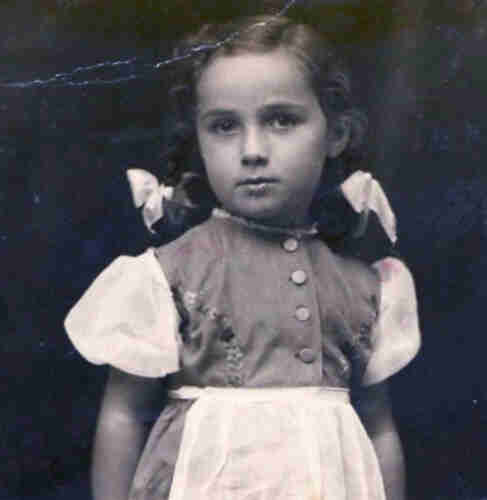 Black and white photograph of a young girl. She is wearing a dress with an apron and frilly short sleeves. There are four buttons in the centre. Her long hair is pinned up in braids. On the braids she has ribbons tied.