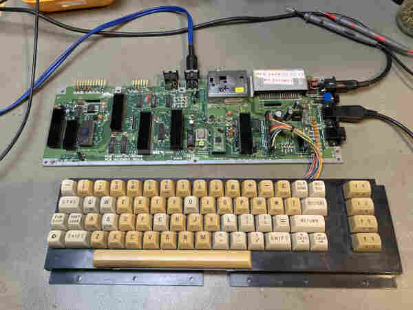 Commodore 64C board on my workbench. The board was recapped and Kim left a little label on the cartridge port cover stating it was done in 2017. A very yellow C64 keyboard is connected to the board for testing. Some multimeter probes are visible on the side.
