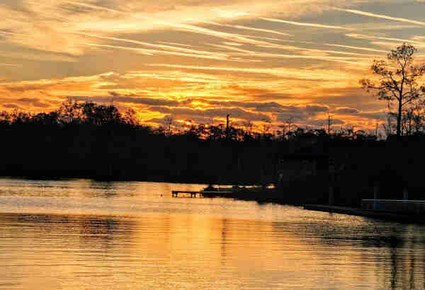 View across a calm body of water against a colorful sunrise over the horizon with shades of yellow and orange reflecting upon the clouds and water.
