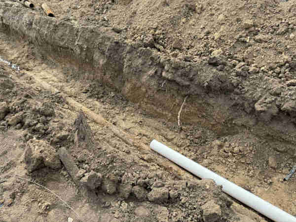 A trench dug into clay soil. A root is visible and a cast iron pipe is in the trench. A segment of white drain pipe is next to it.