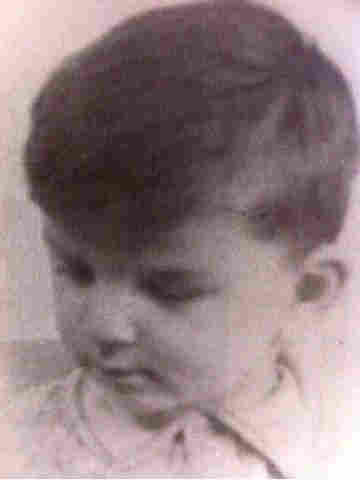 A photo of face and shoulders of a young boy. He is photographed from above and he is looking down. He has short hair and a fringe covering his forehead. He has a collar buttoned under his chin. 