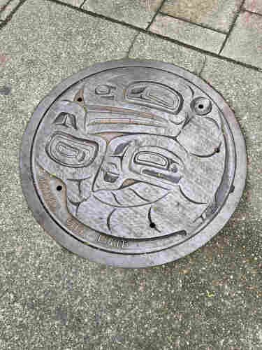 A manhole cover in a sidewalk. It looks like a Haida or Coastal Salish design, with stylized animals. I see at least two heads, maybe a big fish and a small fish, but I'm not entirely sure. It is worn with the passage of many feet. At the bottom are the woods "SEATTLE CITY LIGHT"