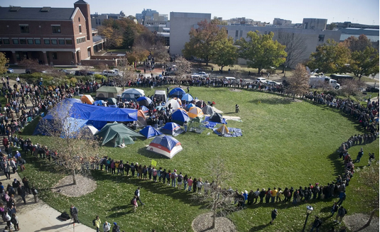 An overhead view of a large encampment, surrounded by a protective circle of people, on college quad. Image from
https://www.nytimes.com/slideshow/2015/11/09/us/students-celebrate-university-of-missouri-presidents-resignation/s/09missouri-campus-ss-slide-VF43.html 