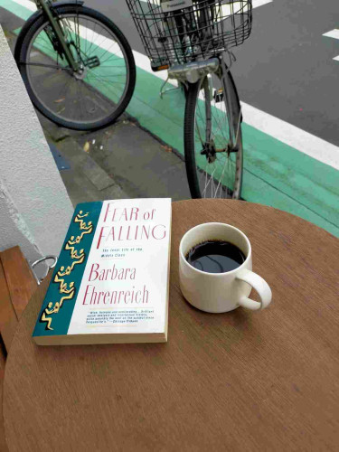 The photo is outside a cafe  On a round wooden brown table is the grey paperback book with a green spine of yellow illustrated men shining the shoes of the men above them, the man at the top holding a martini glass. To the left is a white mug of black coffee. You can see the front of 2 bicycle ls near a green and white strip of paint along the road