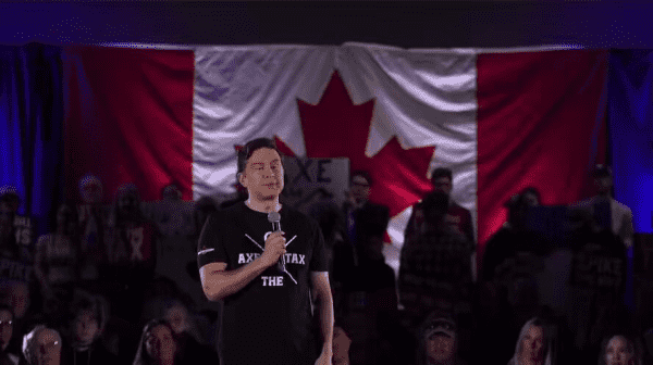 Poilievre speaking at his rally in front of a Canadian flag wearing an "AxeTheTax" t-shirt