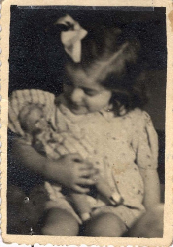A vintage black-and-white photo of a young girl  with a bow in their hair, smiling while holding a small doll. She is sitting.