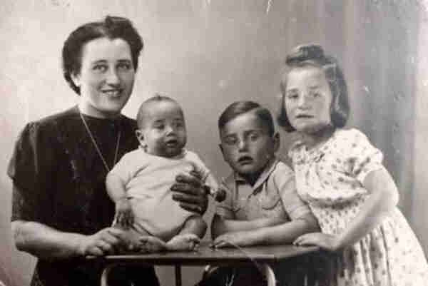 A family picture of a mother and three children. They are all in fron of a table. The mother is on the right. The smallest baby is sitting on the table. His elder brother and sister are standing on the right.